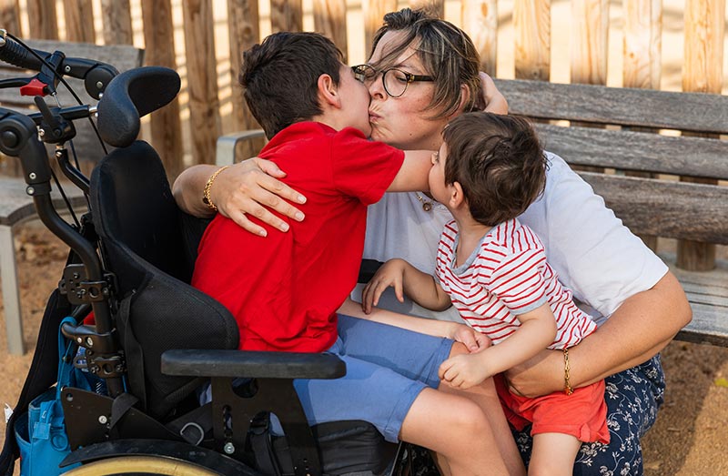 A mother hugs her two young sons, one of whom uses a wheelchair.