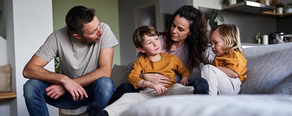 A father, mother and two young children sit together on a sofa.