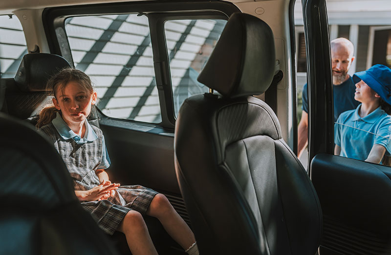 A girl in school uniform sits in the back seat of a car as her family arrives at school.
