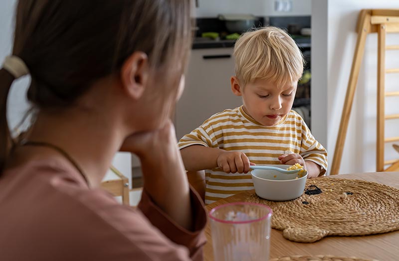 A mother sits at the kitchen table while her toddler eats.
